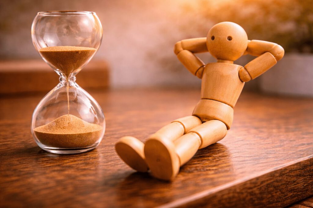 Eine Gliederpuppe sitzt entspannt neben einer Sanduhr in weichem Licht. Symbolbild für die Zeit zwischen den Jahren, in der Führungskräfte zur Ruhe kommen, reflektieren und Kraft für das neue Jahr sammeln.