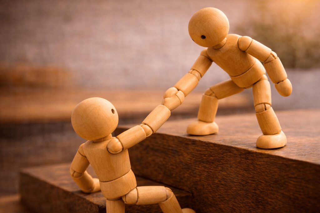 Eine Gliederpuppe hilft einer anderen Figur unterstützend eine Stufe hinauf. Symbolbild für den Karriereanker Dienst und Hingabe, bei dem Werteorientierung, Helfen und sinnstiftende Tätigkeit im Vordergrund stehen.