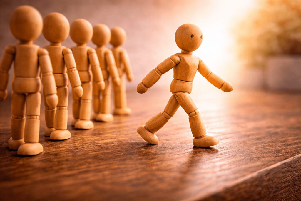 Eine einzelne Figur löst sich aus einer Reihe von anderen Gliederpuppen und geht einen eigenen Weg. Symbolbild für den Karriereanker Selbstständigkeit, das Bedürfnis nach Autonomie, Freiheit und beruflicher Unabhängigkeit.