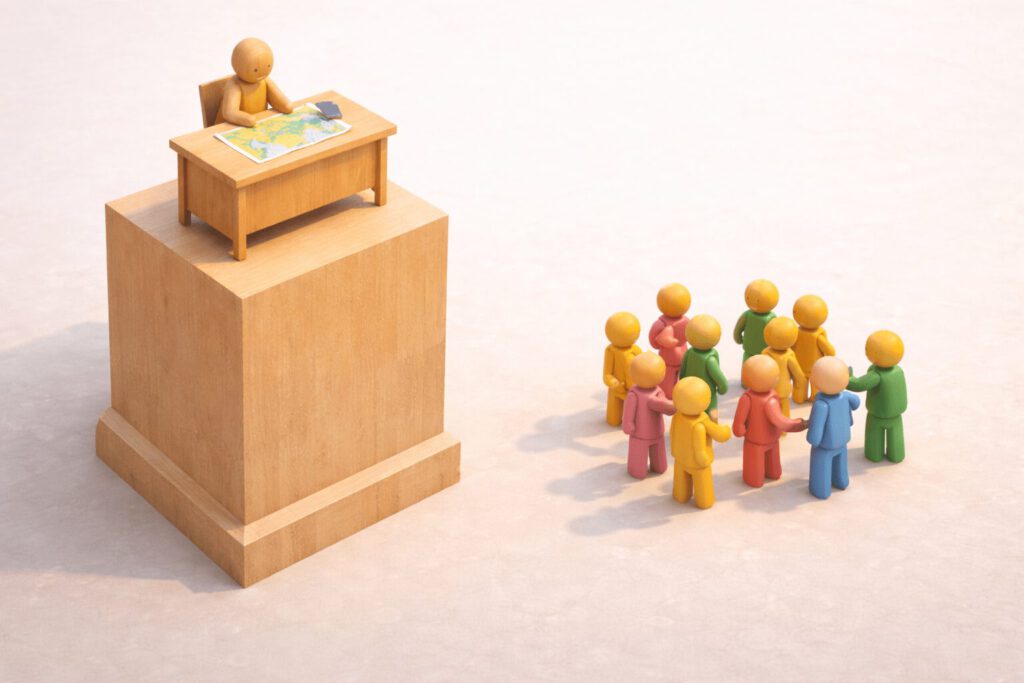 Eine einzelne Holzfigur sitzt isoliert an einem Schreibtisch auf einem sehr hohen Holzpodest. Weit unten am Boden steht eine Gruppe bunter Figuren gesellig im Kreis. Symbolbild für die Einsamkeit an der Spitze und die Distanz zwischen Chef und Team.