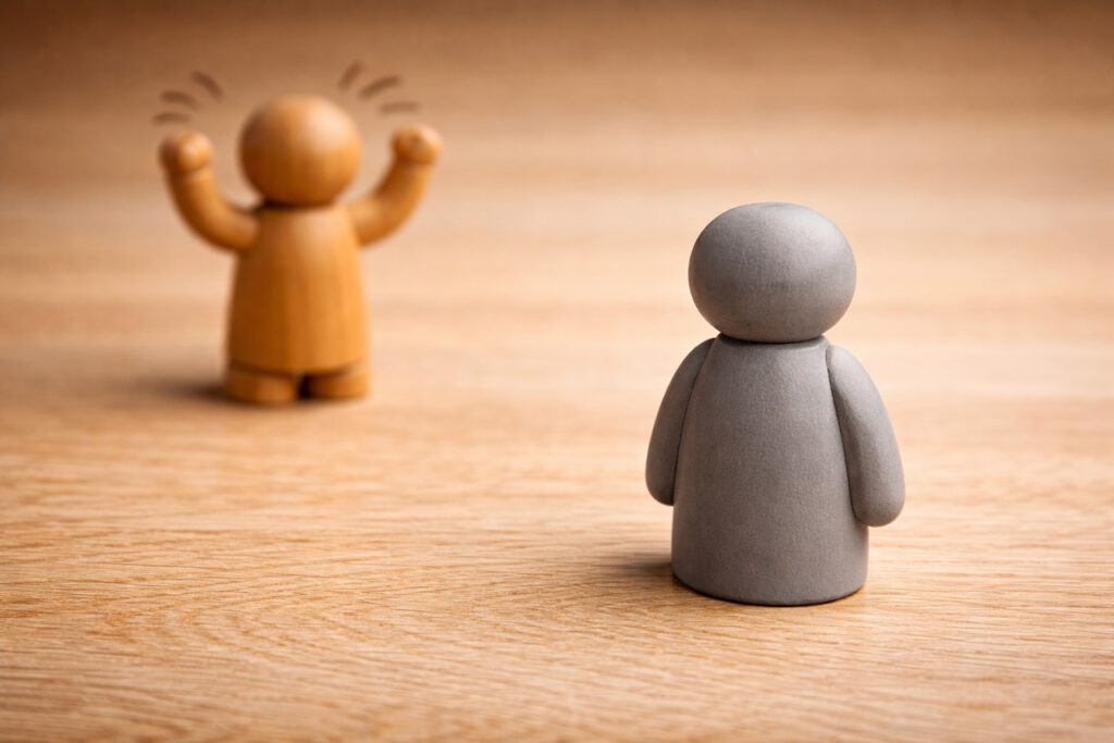 Eine ruhige, graue Holzfigur steht unbewegt im Vordergrund, während im Hintergrund eine andere Figur wild gestikuliert. Symbol für die Grey Rock Methode.